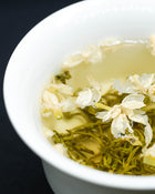 White cup filled with brewed green tea and white jasmine flowers on a dark background