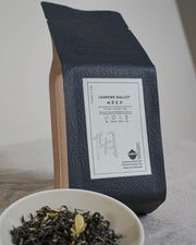Tea packaging with a YIN CHA tea label and a bowl of Chinese jasmine green tea leaves on a neutral background